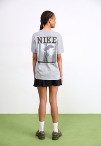 Szara koszulka z czarnym logo Nike na plecach, z graficznym nadrukiem przedstawiającym rzeźbę. Modelka ma na sobie czarną spódnicę. Zielona podłoga.
