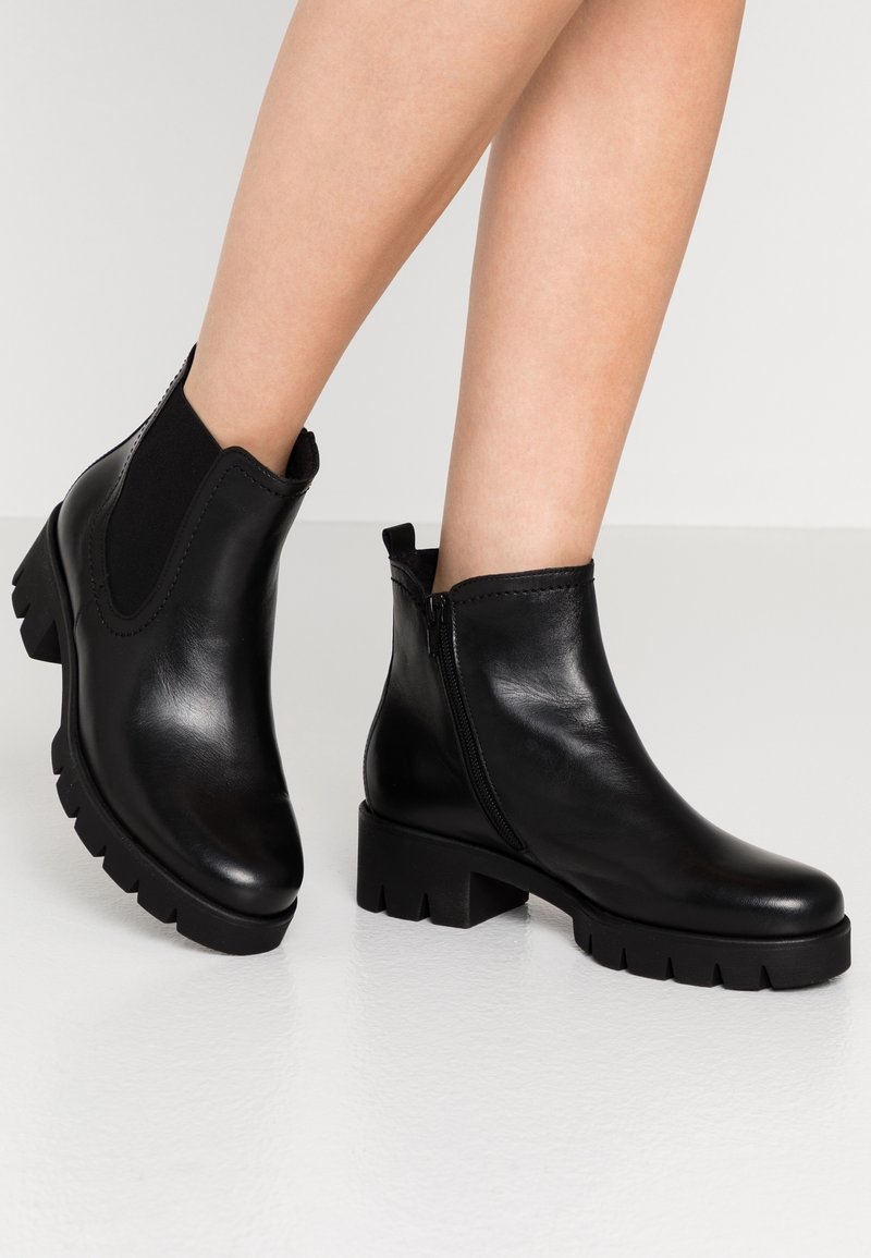 Bottes cheville en cuir noir avec semelles crantées épaisses et fermeture éclair latérale, portées sur jambes nues sur un fond uni.