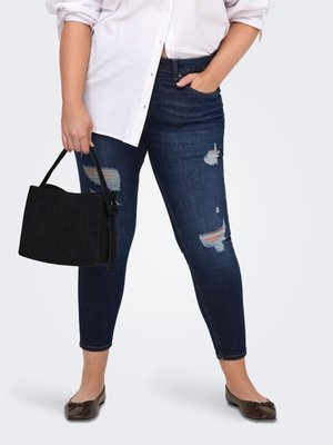 Osoba oblečená v vyhrnutých bielych košeliach, tmavomodrých roztrhaných úzkych džínsoch, hnedých balerínkach, drží čiernu kabelku v ľavej ruke.