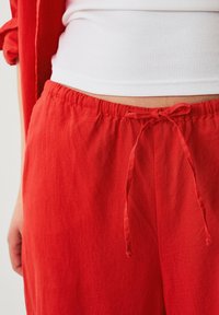 Pantaloni di lino rossi con vita elasticizzata e dettaglio con cordoncino. Il tessuto presenta una superficie leggermente texturizzata. Vista ravvicinata dalla vita in giù.