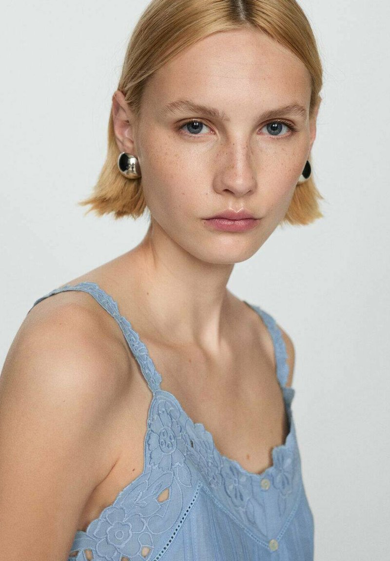 Jasnoniebieska koszulka na ramiączkach z koronkowymi zdobieniami w kwiaty i regulowanymi ramiączkami, o miękkiej fakturze i delikatnych plisach, uzupełniona srebrnymi kolczykami.