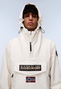 Crème-kleurige hoodie met een voorzak, zwart merkembleem en een Noorse vlagpatch op de onderkant van de voorkant.