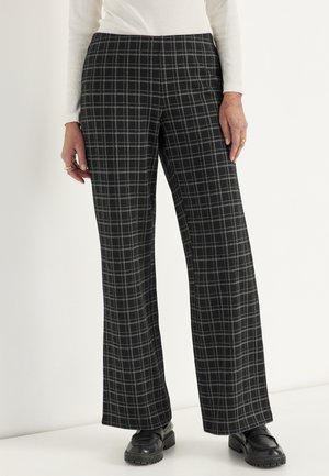 Kvinde iført sort og grå ternede bukser med vide ben, hvid langærmet top og sorte loafers står mod en hvid baggrund.