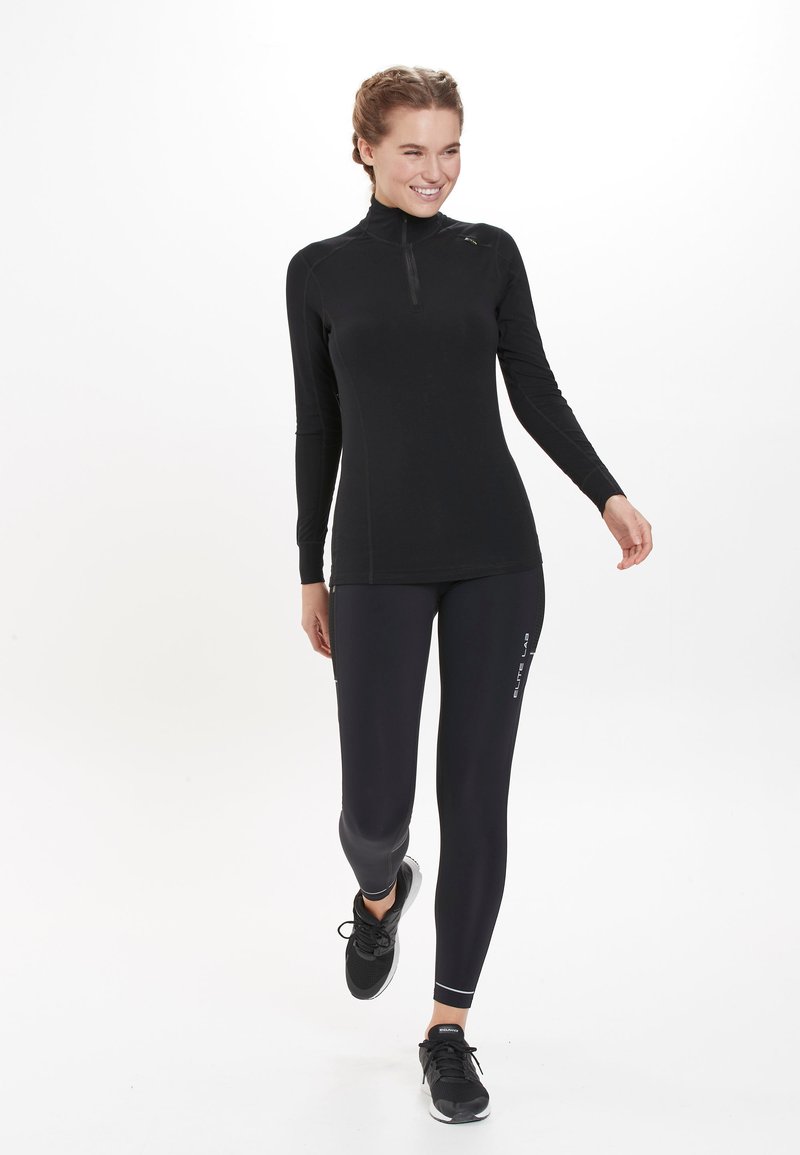 Sort langærmet top med halvlukning og tætsiddende fit, parret med sorte leggings med et subtilt logo og reflekterende detaljer.