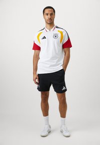 Kortermet hvit polo med røde og gule detaljer, tysk emblem og glidelås krage, kombinert med svarte shorts og hvite joggesko.