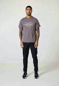 Man with tattoos wears a gray graphic t-shirt, black cargo pants, and black sneakers, standing against a plain light background.