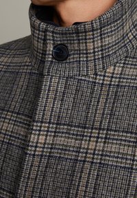 Cappotto di lana con colletto alto, caratterizzato da un motivo a quadri navy, beige e grigio. Un bottone nero accentua il lato destro del colletto.
