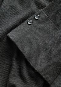 Tissu en maille gris foncé avec deux boutons ronds foncés cousus près d'un bord plié, montrant une couture fine et texturée.