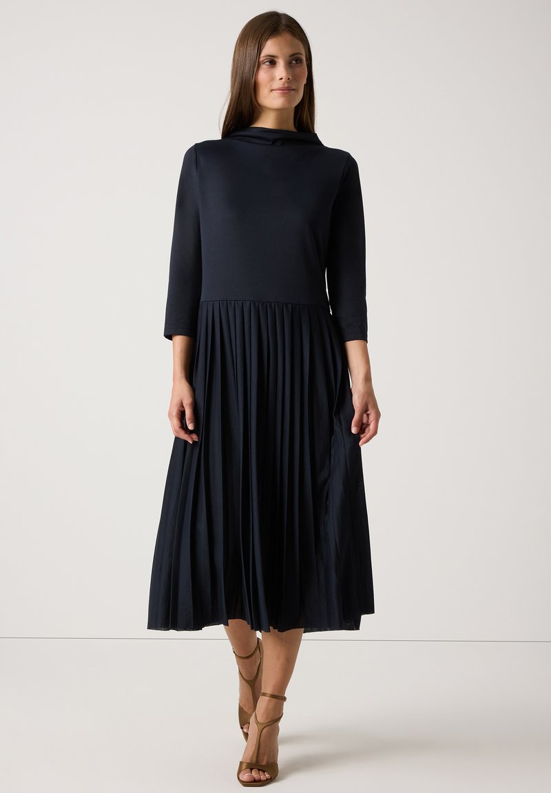 Granatowa sukienka z rękawami o długości trzy czwarte i plisowanym dołem, z dekoltem typu cowl oraz gładką teksturą. Do kompletu pasują sandały na obcasie w odcieniu beżowym.