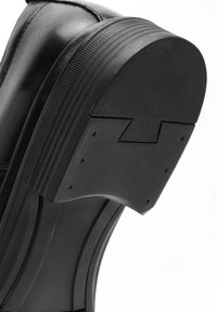 Zwarte leren schoen met een gestructureerde rubberen zool, voorzien van een gelaagd ontwerp en een gebogen hak met een opvallende schuine snit.