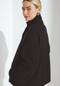 Sort oversized jakke med høj krave, lavet af tekstureret stof, med dropped shoulders og en ren silhuet. Sølvøreringe er synlige.