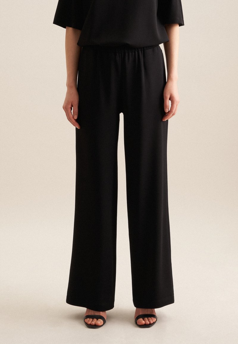 Pantalons larges noirs en tissu lisse, avec une taille élastique. L'ourlet inférieur a une finition soignée. Portés avec des sandales à talons noires.