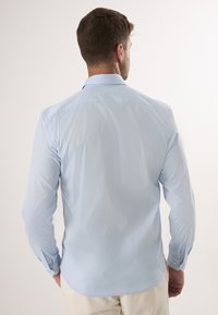 Helle blaue Langarmshirt, glattes Material, Knopfmanschetten, klassischer Kragen, abgerundeter Saum und dezente Stichdetails.