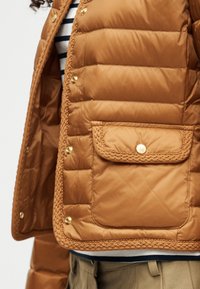 Καφέ μπουφάν με επένδυση καπιτονέ, μεγάλο μπροστινό τσεπάκι και χρυσά κουμπιά με τσιμπιδάκι, φορεμένο πάνω από ριγέ πουκάμισο και μπεζ παντελόνι.