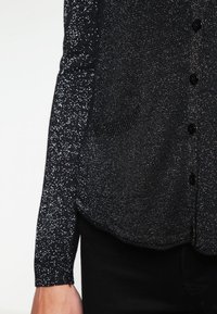 Svart glittrig cardigan med strukturerad yta, med knappstängning framtill och två fickor, tillverkad av ett mjukt, elastiskt material.