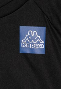 Melna auduma, kas rotāta ar zilu taisnstūra logotipu, divām baltām figūrām un vārdu "Kappa" baltā krāsā, centrāli izvietots.