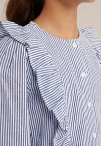 Mėlynos ir baltos juostelės marškiniai su raukinėtą apykakle, mygtukų uždarymu ir tekstūruotu audiniu, akcentuojančiu vertikalias juosteles ir švelnias kraštines.