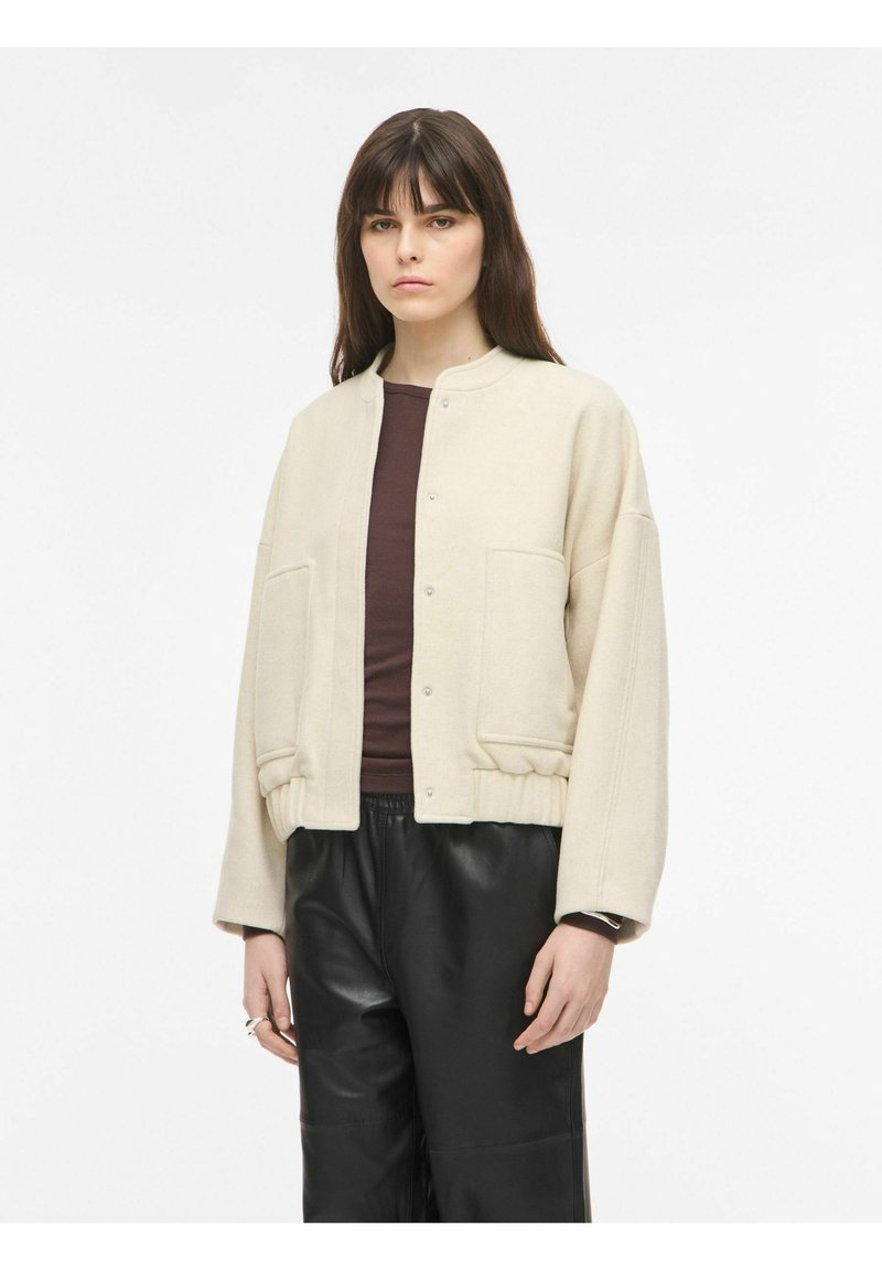 Jeune femme aux cheveux bruns raides porte une veste beige boutonnée, un haut marron foncé et un pantalon en cuir noir sur un fond blanc uni.