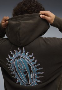 Άτομο με σκούρα κουκούλα σηκώνει την κουκούλα, με μεγάλο μπλε και μοβ ηλιοειδές γραφικό και στυλιζαρισμένο κείμενο στην πλάτη.