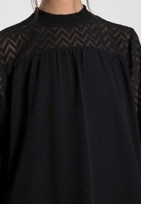 Haut noir présentant un motif en zigzag texturé et un accent en dentelle transparente au niveau de l'empiècement, avec un design plissé et un col montant smocké.