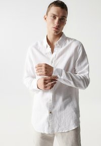 Camisa blanca de manga larga hecha de lino con cuello abotonado, que presenta una textura sutil y un único bolsillo en el pecho.