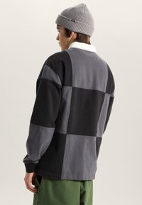 Ung person som bär en grå mössa och en svart-grå lapptäckströja med lång ärm, vänd åt sidan med gröna byxor mot en enfärgad bakgrund.