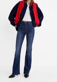 Veste en coton bleu marine avec un gilet en laine rouge, un débardeur blanc et un jean flare bleu foncé. Tissu texturé, silhouette ajustée et escarpins noirs pointus.