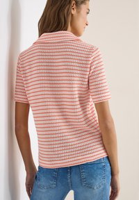 Femme de dos portant un haut en tricot à manches courtes blanc avec des rayures horizontales orange, associé à un jean en denim bleu.