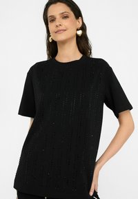 Schwarzes T-Shirt mit kurzen Ärmeln, das vertikale Reihen aus schwarzen Pailletten besitzt, einen runden Ausschnitt hat und aus weichem Baumwollmaterial mit lockerer Passform gefertigt ist.