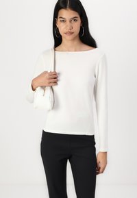White, long-sleeve top in lightweight fabric with a round neckline, paired with black pants. Model holds a small white handbag.