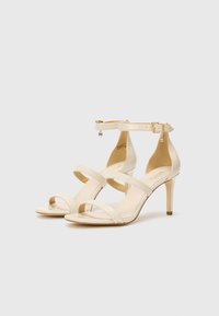 Sandali con tacco alto in vernice beige, dotati di un design a tre cinturini e fibbia alla caviglia, con un delicato charm dorato sul cinturino alla caviglia.