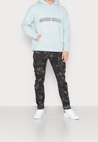 Světle modrá mikina s kapucí s nápisem "MODERN REALITY", doplněná o černé maskáčové otevírací kalhoty a bílé tenisky. Má přední kapsu na břiše.