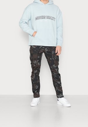 Felpa con cappuccio azzurra chiara con la scritta "MODERN REALITY", abbinata a pantaloni cargo nero mimetici e sneakers bianche. Presenta una tasca a marsupio frontale.