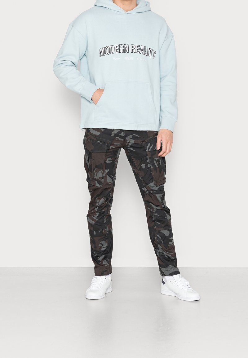Světle modrá mikina s kapucí s nápisem "MODERN REALITY", doplněná o černé maskáčové otevírací kalhoty a bílé tenisky. Má přední kapsu na břiše.
