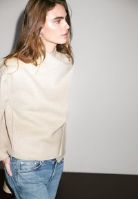 Pull en laine beige à col montant, associé à un jean en denim bleu clair. Texture douce, design minimaliste et coupe décontractée.