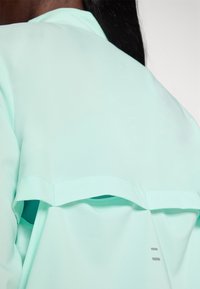 En mintgrön lättviktsöverdel med ett lagerdesign, slät textur, subtila sidöppningar och minimalistiska grå accenter.