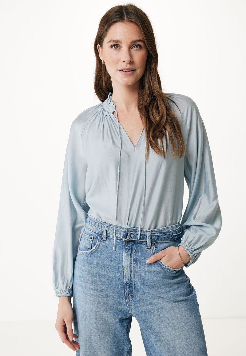 Blusa azzurro chiaro con scollatura a V e dettaglio arricciato sul collo, maniche lunghe e polsini elasticizzati, abbinata a jeans in denim a vita alta.
