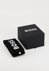 Pochette en daim noire avec le logo "BOSS" à côté d'une boîte noire arborant le même logo. Les deux articles sont placés sur un fond clair.