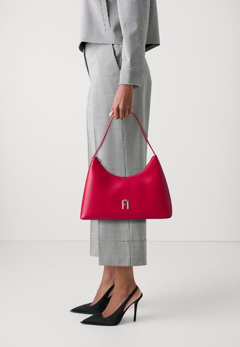 Sac en cuir rouge au design incurvé avec des accessoires en argent, tenu par une personne vêtue d'un ensemble pied-de-poule noir et blanc et de talons.