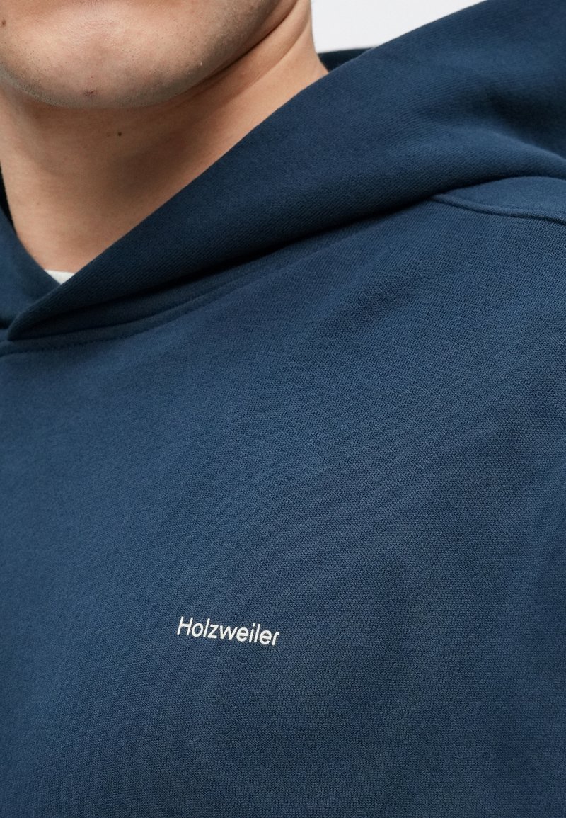 Navy blue hoodie made of cotton, featuring a large hood and a white "Holzweiler" logo on the chest. Smooth texture, casual design.