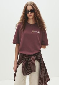 Ung kvinde med langt krøllet hår iført mørke solbriller, en bordeaux t-shirt med tekst, lyse bukser og en stribet skjorte bundet om taljen.