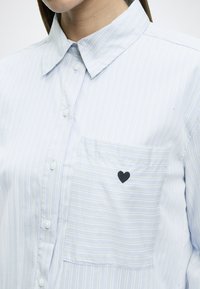 Ανοιχτό μπλε μακρυμάνικο ριγέ πουκάμισο με λευκές και κίτρινες γραμμές, κουμπιά στο μπροστινό μέρος και μικρή μαύρη κεντημένη καρδιά στην τσέπη στο στήθος.