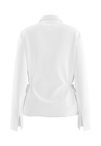 Chemise blanche à manches longues avec col, taille ajustée avec liens latéraux et larges poignets fendus, vue de dos sur un fond uni.