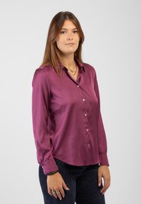 Camicia di seta magenta con maniche lunghe, chiusura a bottoni e colletto. Tessuto liscio con un design leggermente aderente. Abbinata a pantaloni in velluto a coste scuri.