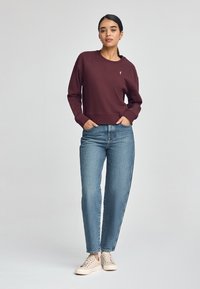 Felpa bordeaux con colletto rotondo e polsini a costine, abbinata a jeans blu a vita alta e sneakers beige chiaro. Dettaglio semplice del logo.