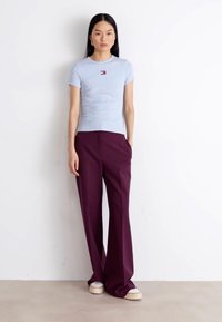 T-shirt aderente azzurro chiaro con un piccolo logo, abbinato a pantaloni ampi bordeaux e scarpe beige, in piedi su uno sfondo neutro.