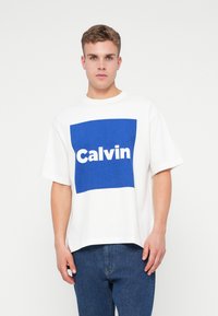 Młody mężczyzna ubrany w białą, dużą koszulkę z dużym niebieskim kwadratem i napisem "Calvin", w połączeniu z niebieskimi dżinsami, stojący na tle jednolitym.