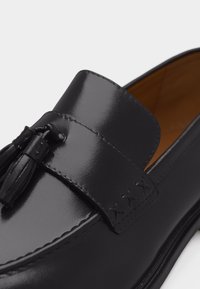 Mocassin à pampilles en cuir noir avec une bande cousue sur le dessus, présentant une couture détaillée près du bord avant.