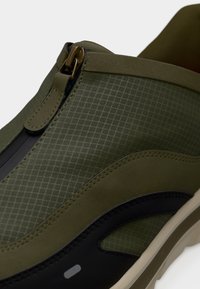 Πράσινα χακί παπούτσια τύπου slip-on με υφή στην επάνω επιφάνεια, που διαθέτουν μαύρη λεπτομέρεια, χρυστό φερμουάρ και εξωτερική σόλα σε κρεμ απόχρωση με σχεδιασμένο μοτίβο.