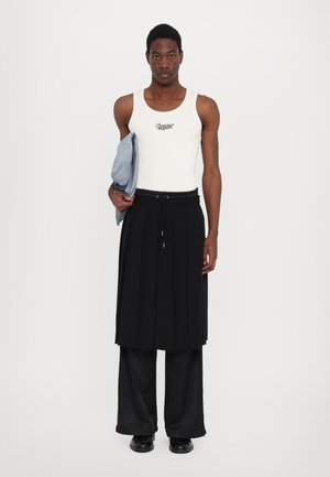 White tank top with black lettering, layered over a black, pleated skirt and wide-leg black pants, holding a light blue denim jacket.